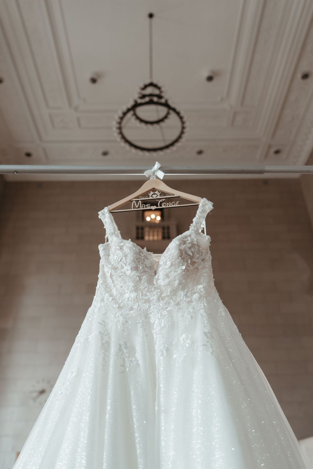 White lace A-line wedding dress hanging on personalized wooden hanger in elegant bridal suite.
