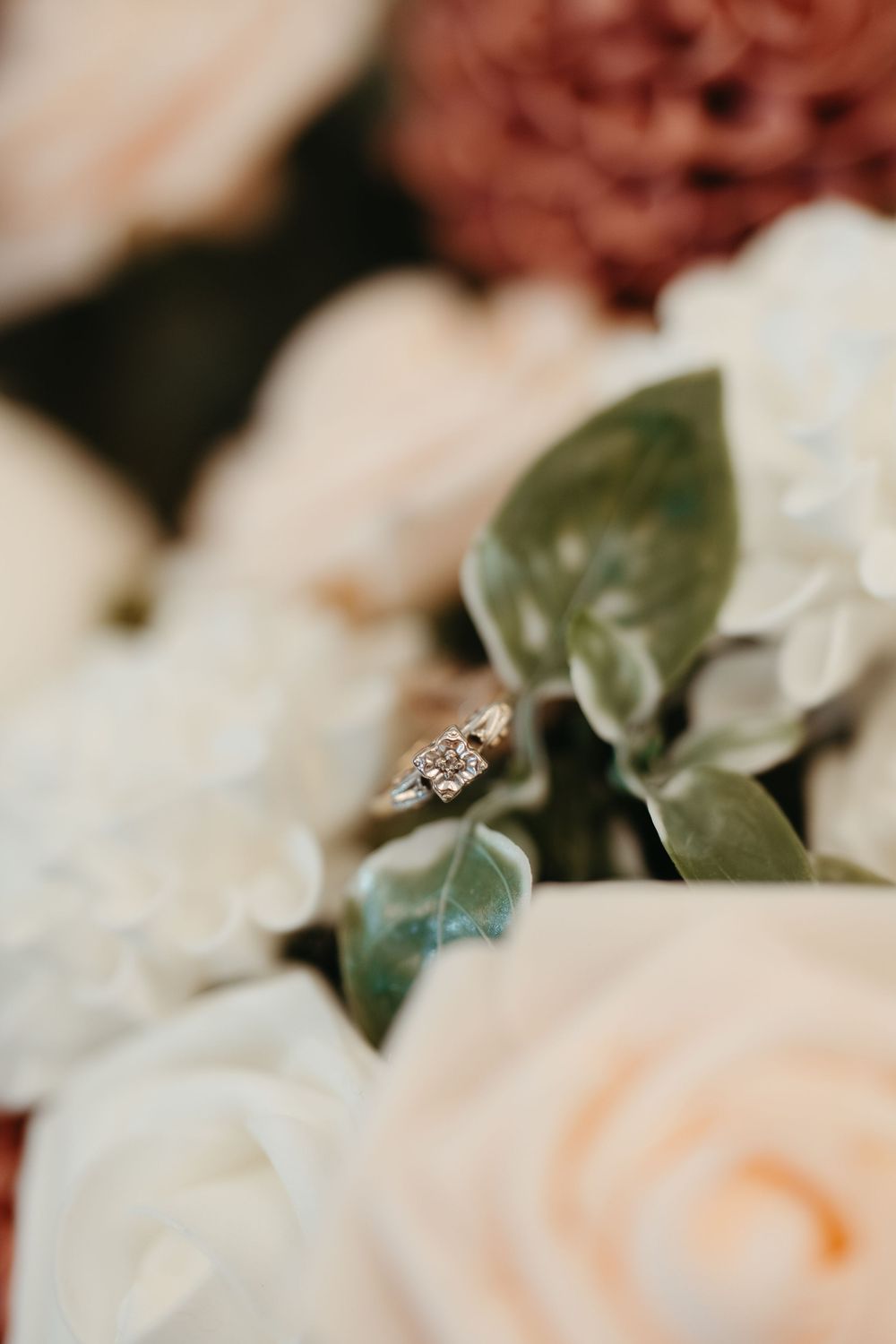 Diamond engagement ring resting on green leaves among soft white and blush pink roses and dusty mauve flowers in a bridal bouquet.
