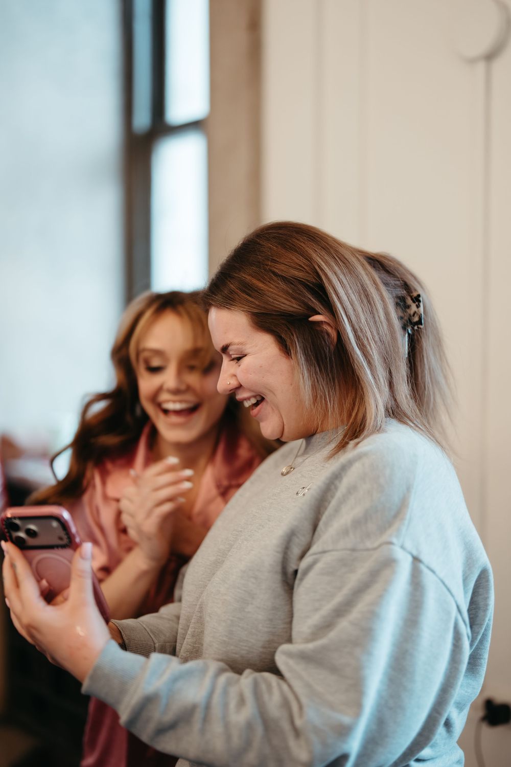 Two women laughing together while looking at a smartphone, sharing a joyful moment indoors.