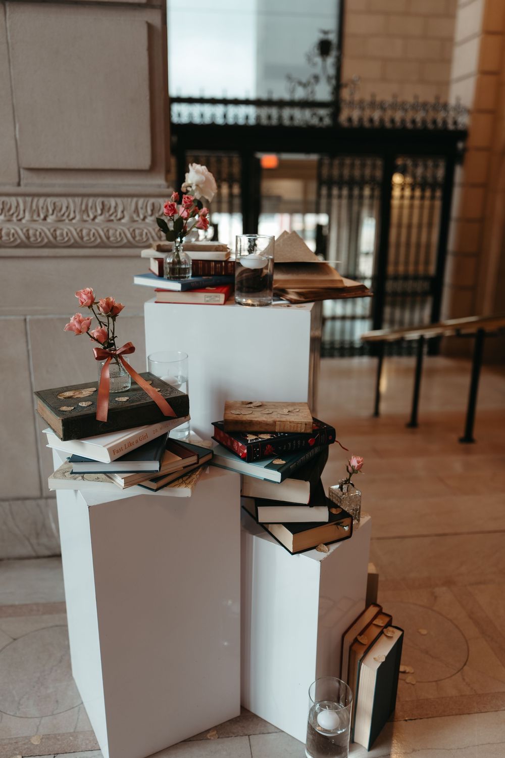 Elegant book display on white pedestals with pink flowers and candles, perfect for a literary-themed wedding decor.