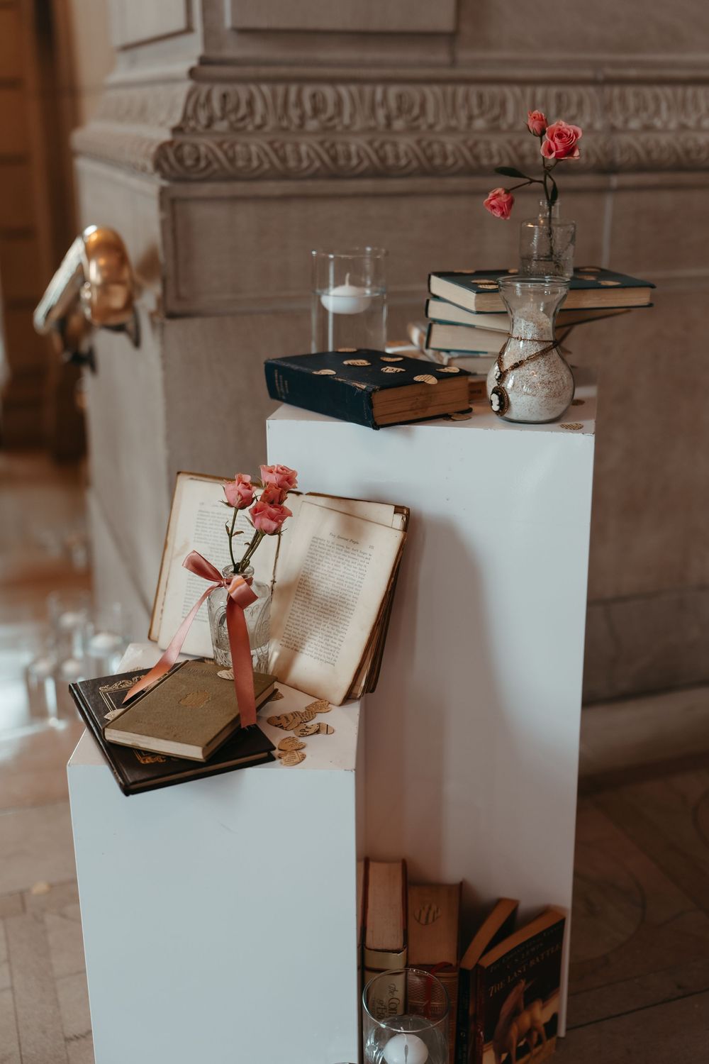Elegant book display on white pedestals with pink roses, candles, and vintage books in a classic stone interior.