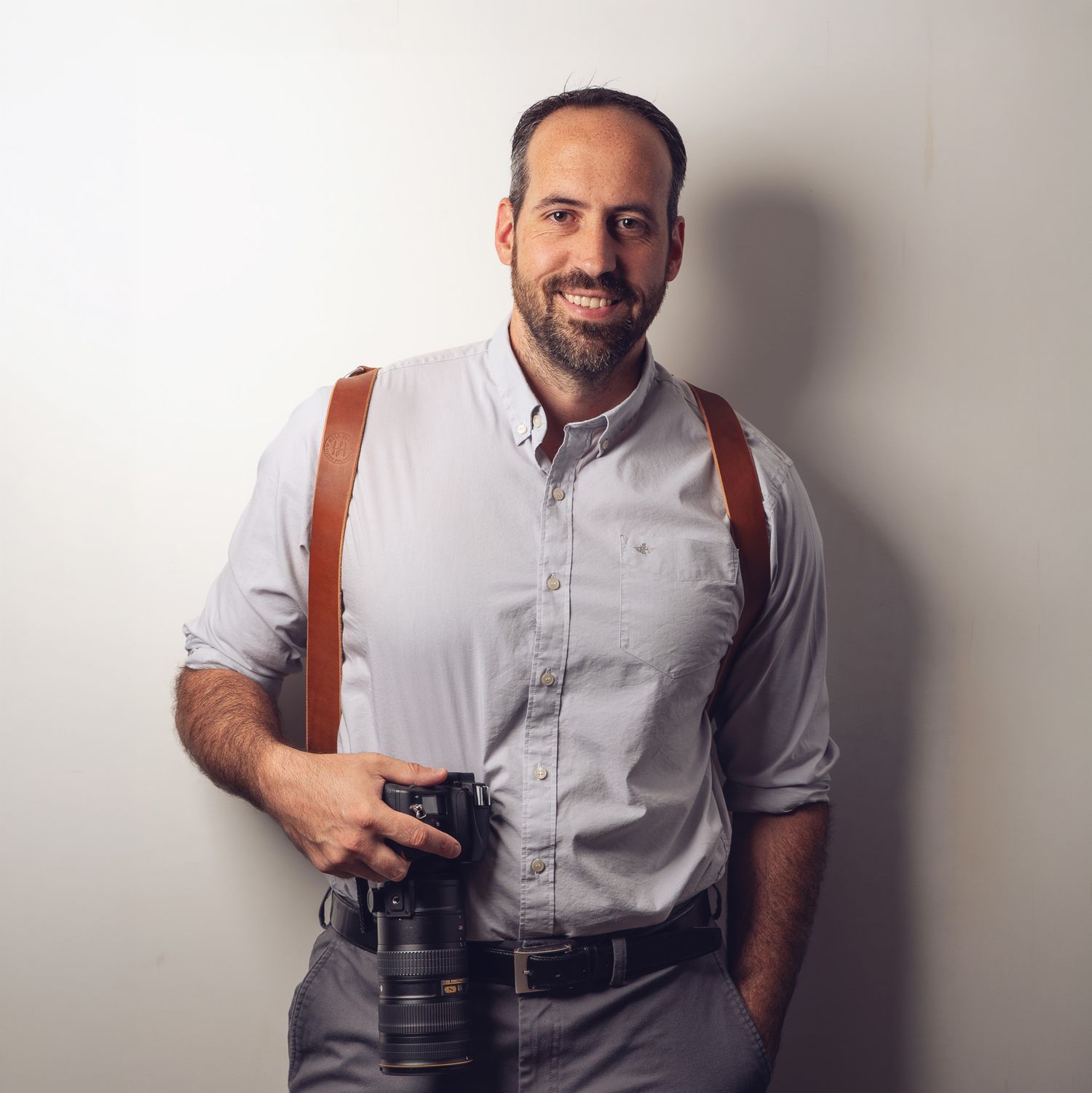 Smiling male photographer in gray shirt with brown leather camera strap holding DSLR camera, posing against light wall.