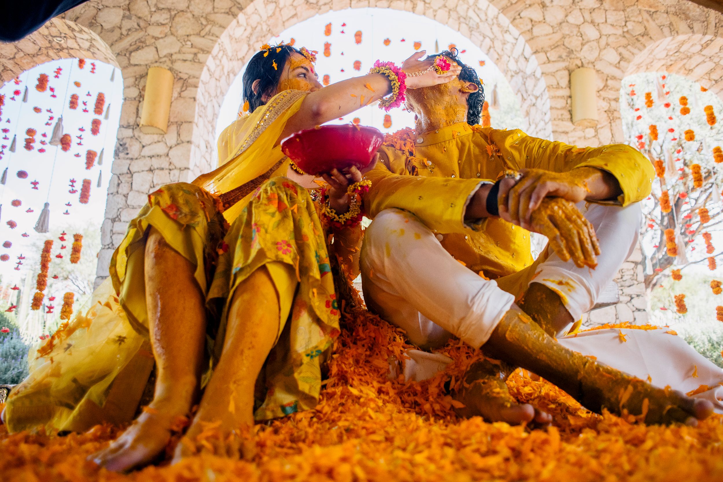 Couple in yellow outfits celebrating Holi festival, covered in orange marigold flowers and colorful powder.