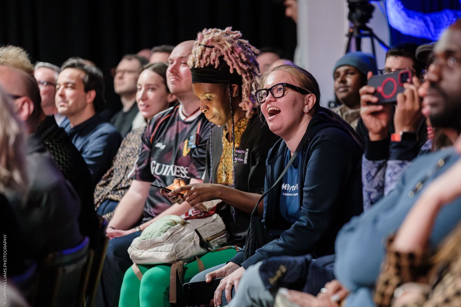 Diverse audience seated at an indoor event, smiling and engaged, with colorful outfits and phones capturing the moment.