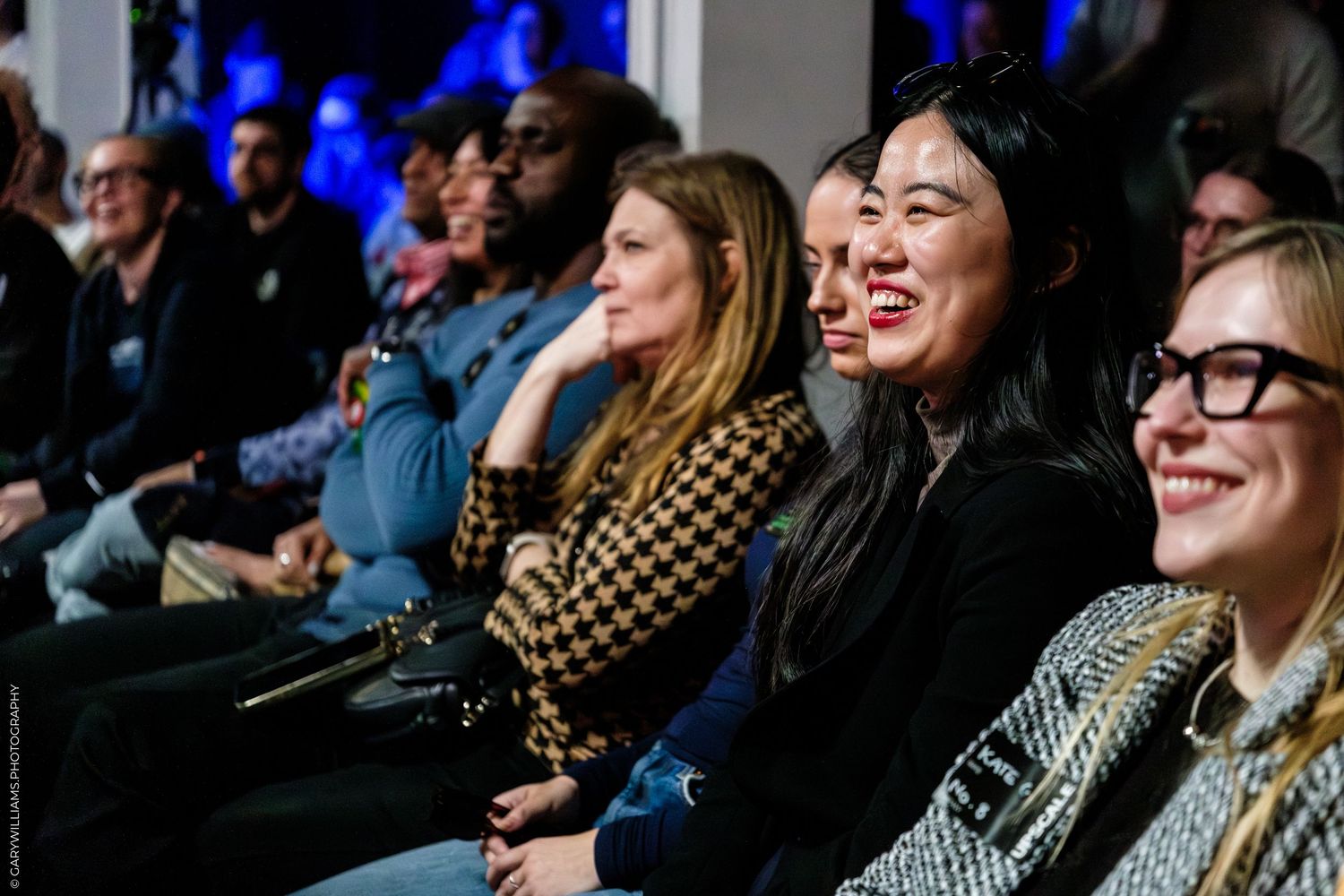 Diverse audience laughing and smiling at an indoor event, seated in rows under blue stage lighting.
