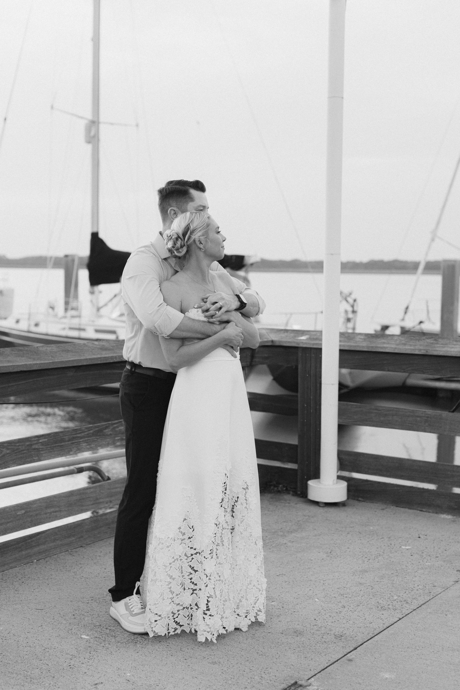 Black and white photo of a couple embracing on a marina dock, bride in lace gown, sailboats in background.