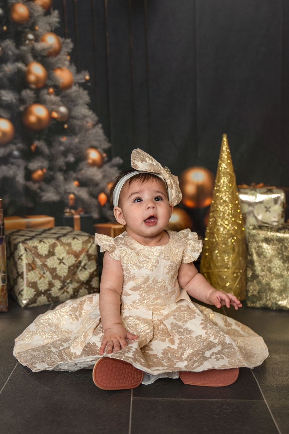 Baby girl in gold lace dress and bow headband sits before Christmas tree with golden ornaments and gifts.