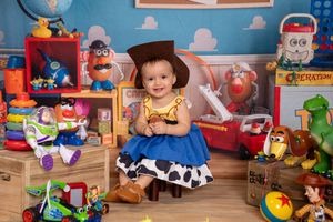 Toddler dressed as Woody from Toy Story sits surrounded by colorful toys and Toy Story characters in a themed photo shoot.