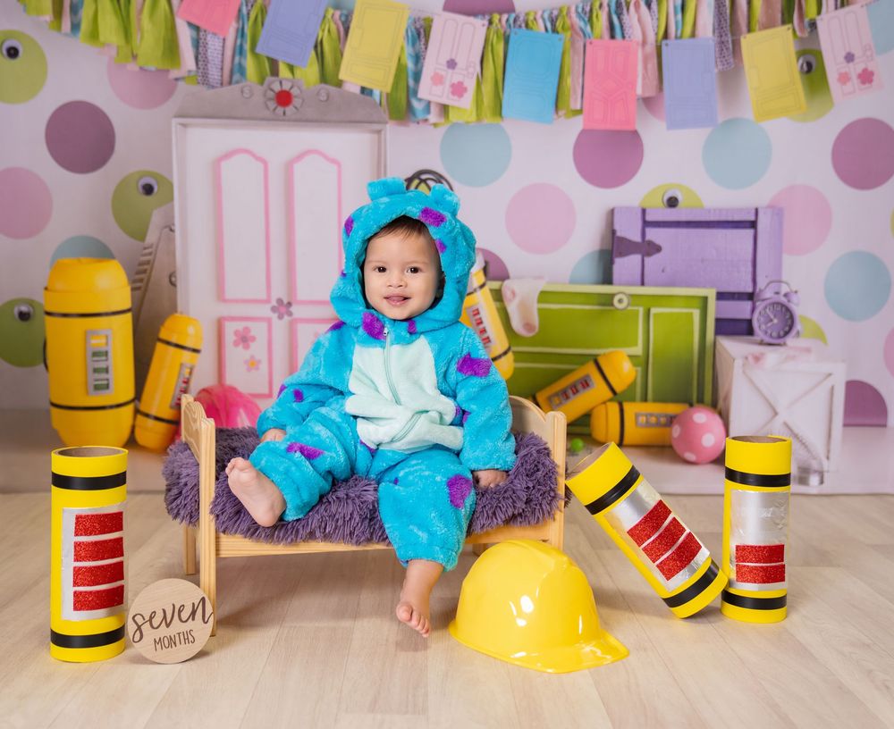 Baby in blue monster costume sits on small bed surrounded by colorful props in a playful photo studio setting.
