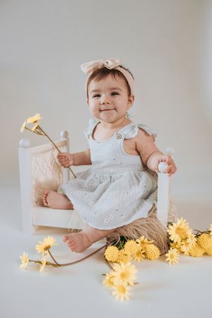 Smiling baby girl in white dress and bow headband sitting in miniature chair surrounded by yellow flowers.
