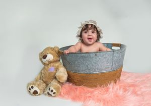 A happy baby sits in a rustic metal tub beside a teddy bear on a pink fur rug against a light background.