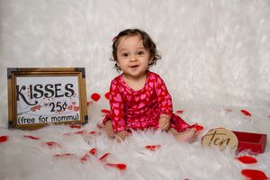 Baby girl in red heart outfit sits on white fur with rose petals and a Kisses 25 cents Valentine sign.