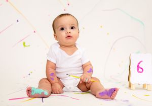A cute baby in a white onesie sits surrounded by colorful paint marks, celebrating their 6-month milestone.