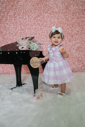 Smiling baby girl in pink plaid dress stands beside mini grand piano at her eleven month milestone photo shoot.