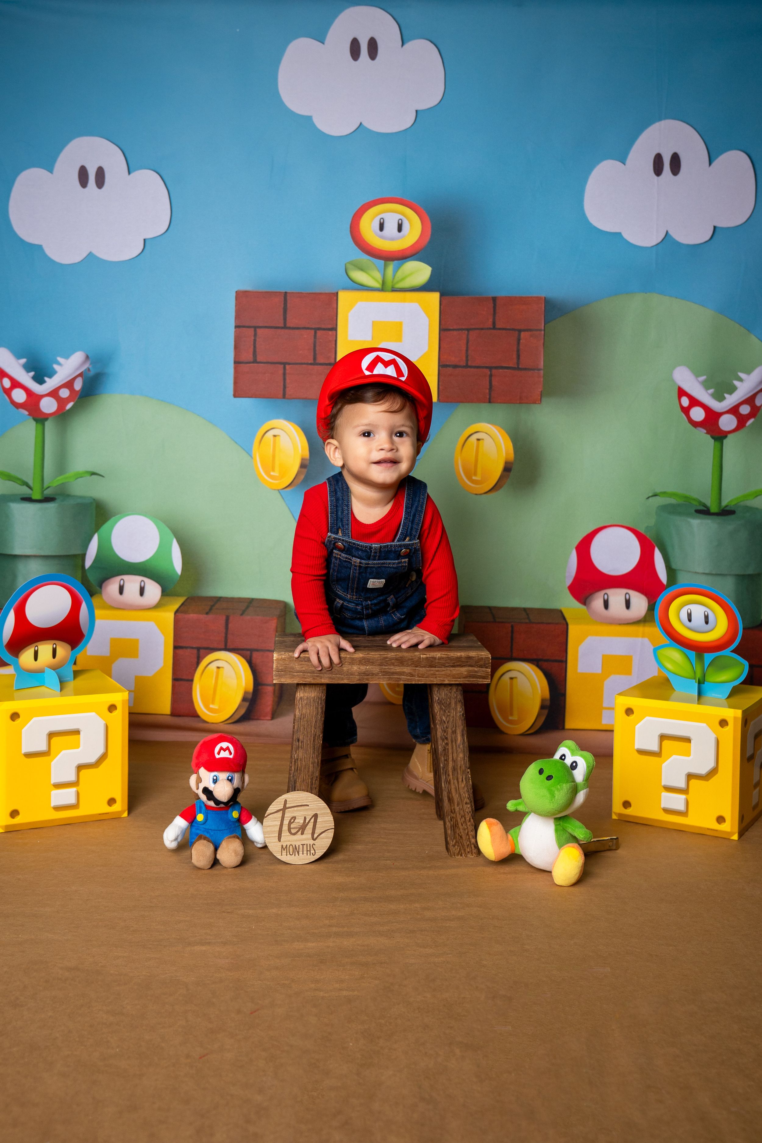 Toddler boy in Mario costume sits among Super Mario Bros themed decorations for a first birthday photo.