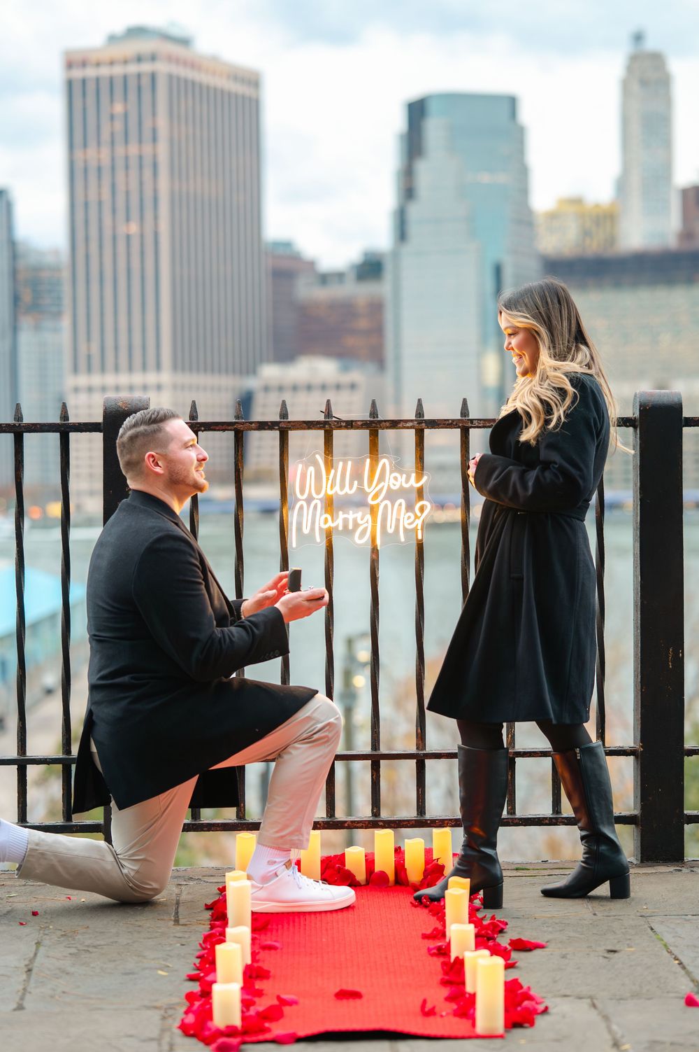 Man proposing on one knee with ring box, red carpet, candles, and Will You Marry Me sign, NYC skyline backdrop.