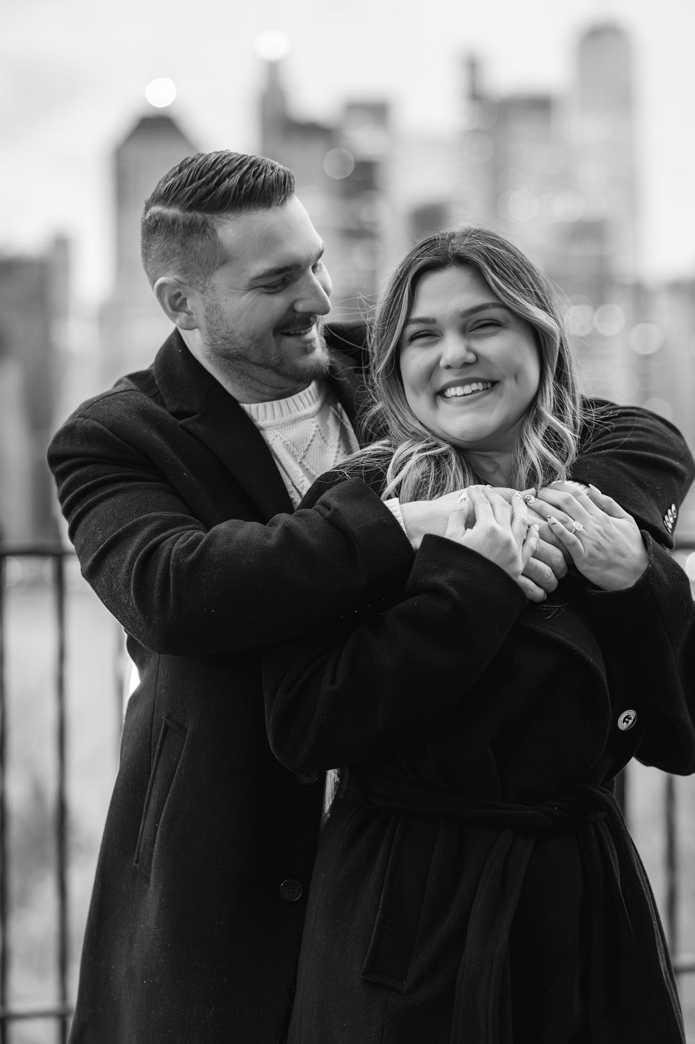 Black and white photo of a smiling couple embracing on a rooftop with a city skyline backdrop.
