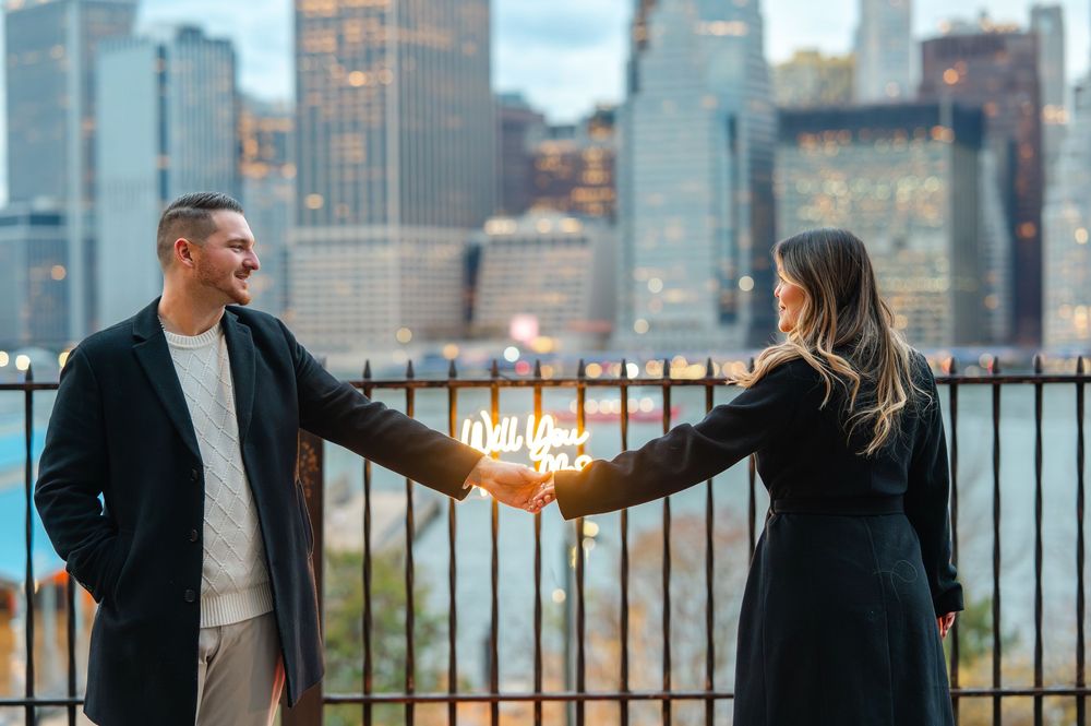 Couple holding a Will You Marry Me neon sign with NYC skyline backdrop at dusk.
