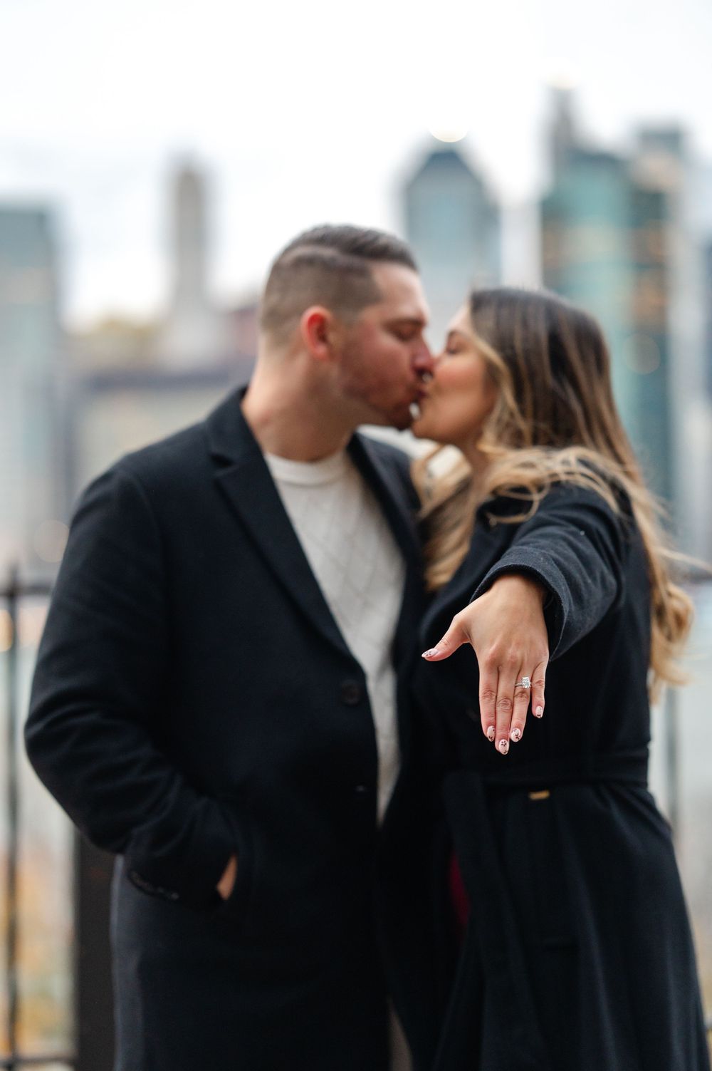 Couple sharing a kiss on a rooftop while the woman extends her hand to show off her engagement ring with a city skyline backdrop.