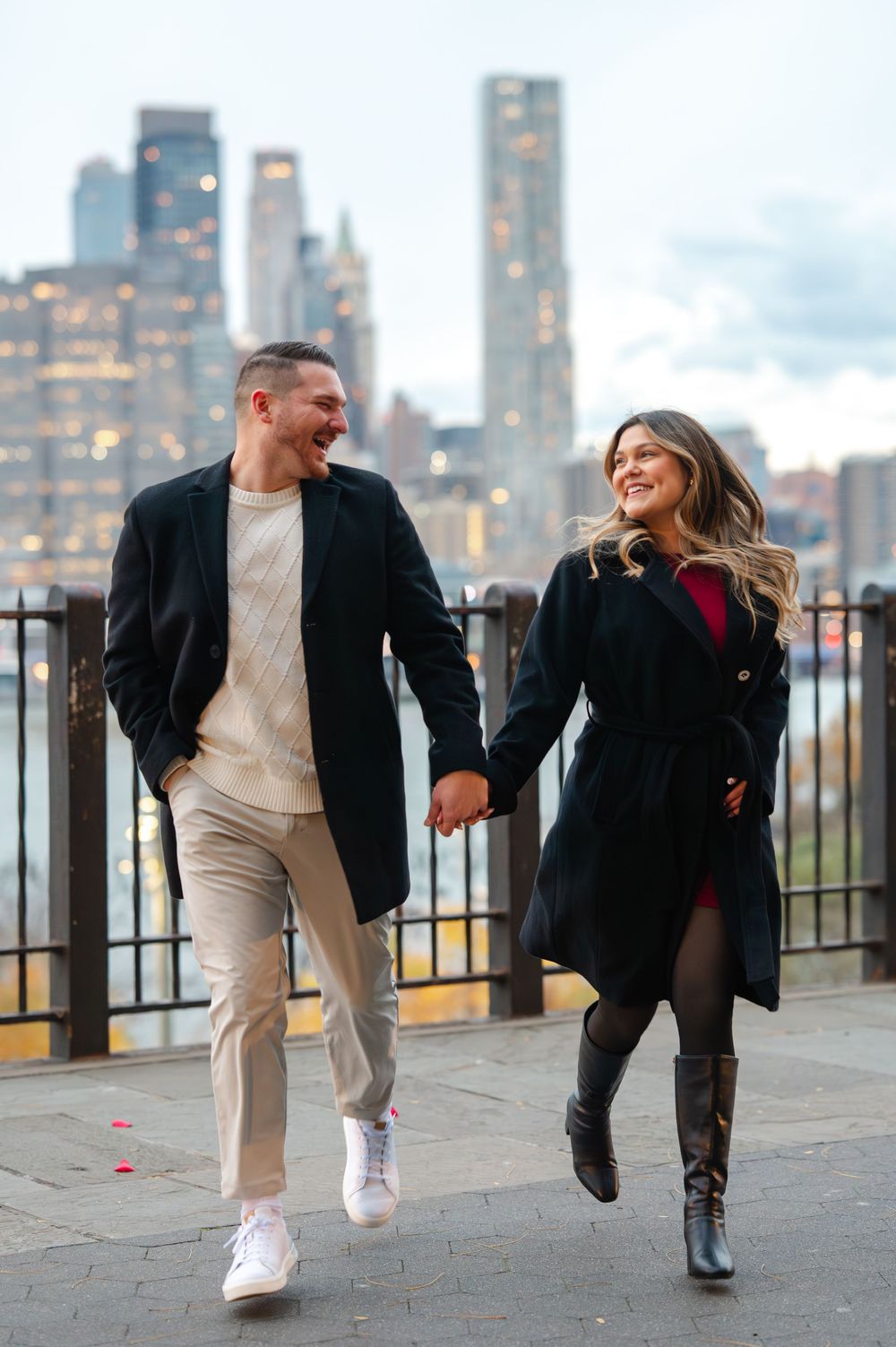 Couple holding hands and laughing while walking along NYC waterfront promenade with Manhattan skyline at dusk.