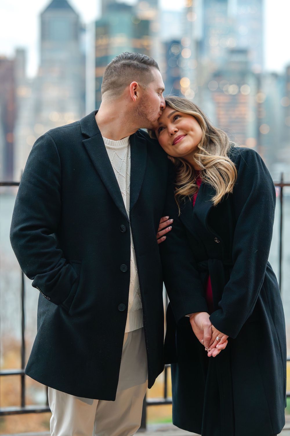 Couple sharing a romantic kiss with NYC skyline backdrop, both wearing elegant black coats on a winter day.