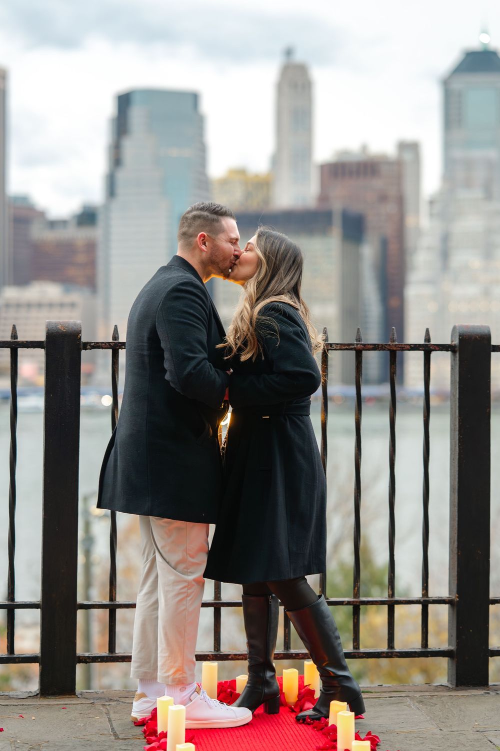 Couple kissing during a romantic proposal on a red carpet with candles, NYC skyline backdrop.