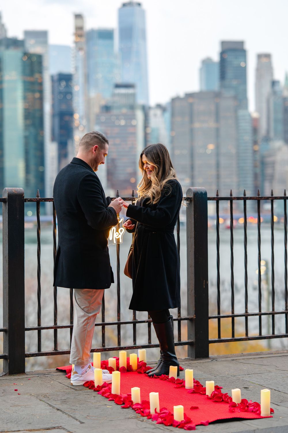 Romantic marriage proposal on NYC waterfront with red carpet, candles, and Manhattan skyline backdrop at dusk.