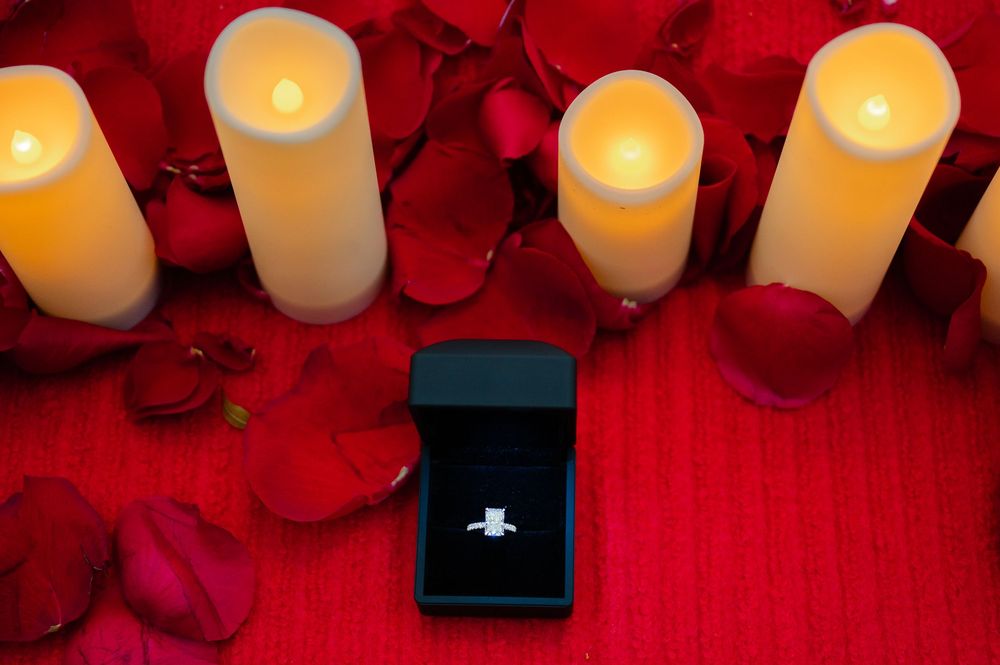 Diamond engagement ring in black box surrounded by red rose petals and glowing candles on red background.