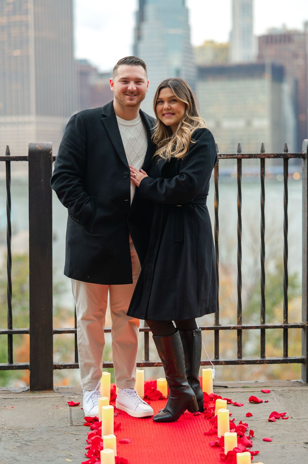 Couple poses on red carpet with candles and rose petals by NYC waterfront railing during romantic proposal setup.