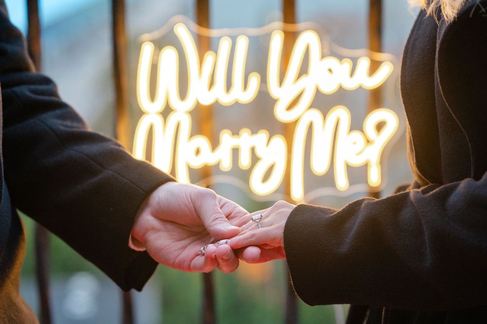 Person holding a ring during a marriage proposal in front of a glowing neon Will You Marry Me sign.