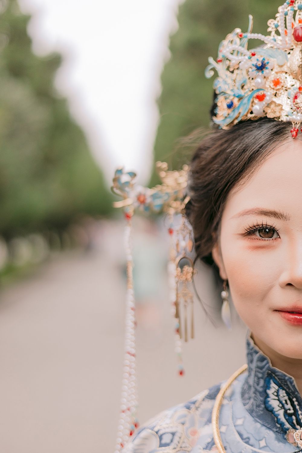 Young woman in traditional Chinese bridal attire with ornate floral crown and dangling gold hair accessories outdoors.