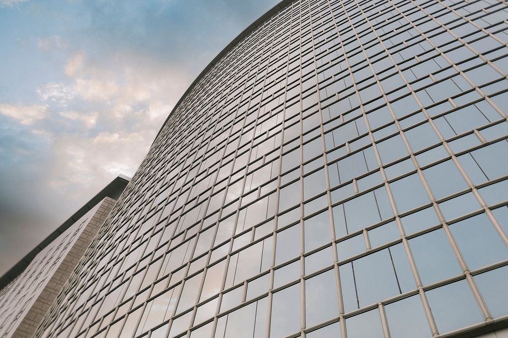 Modern glass skyscraper facade curving upward against a cloudy blue sky, showcasing reflective windows in a grid pattern.