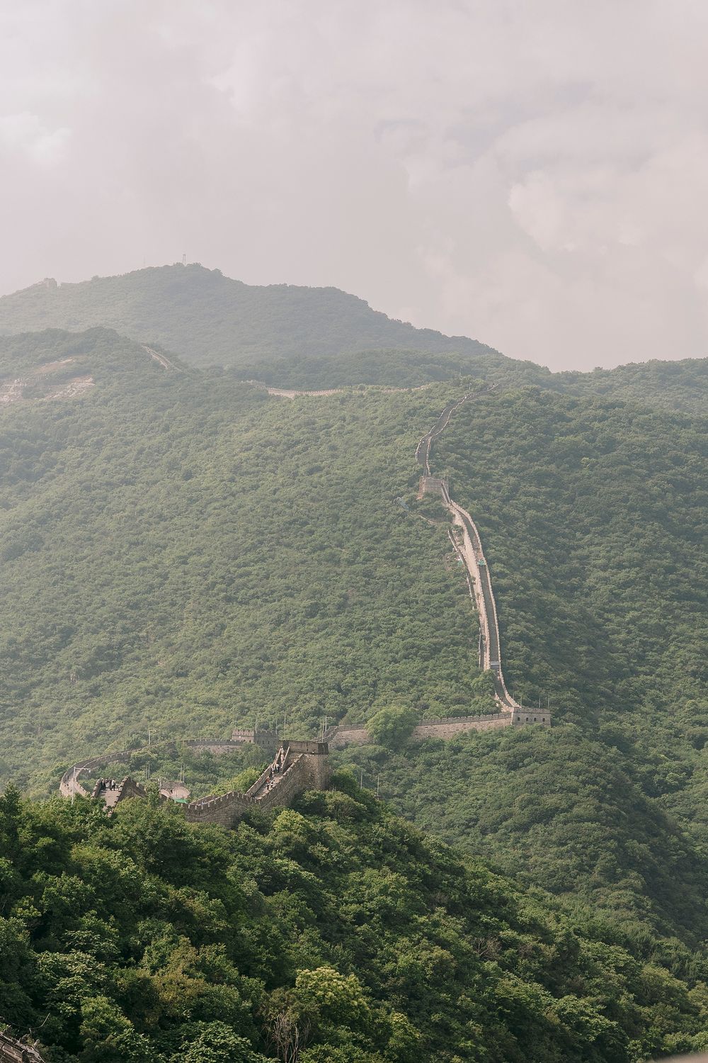 The Great Wall of China winds through lush green mountains under a cloudy sky.