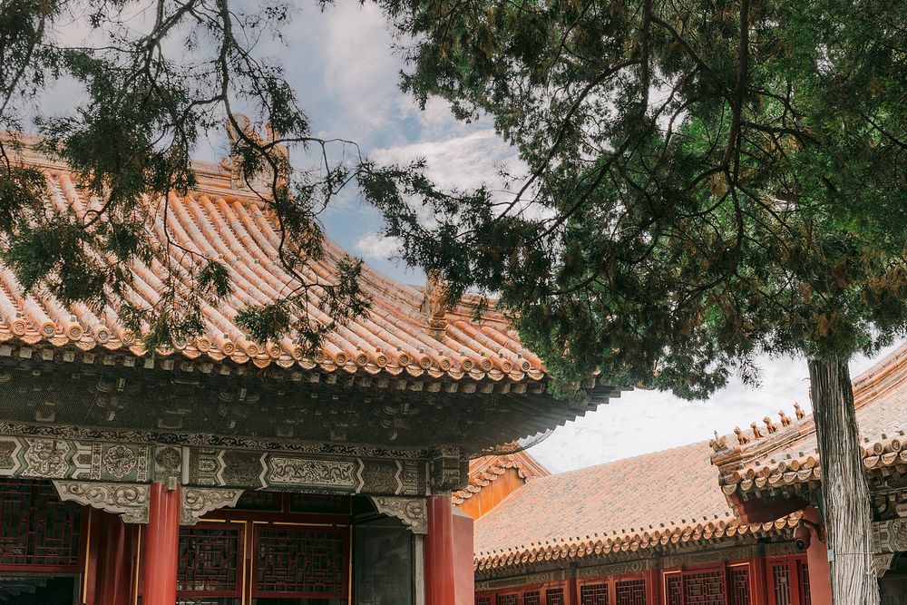 Traditional Chinese architecture with ornate red columns, decorative tiled rooftops, and lush pine trees under a partly cloudy sky.