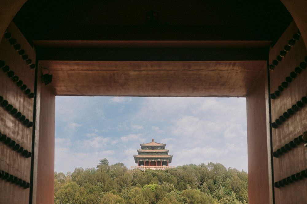 A traditional Chinese pagoda rises above lush green trees, framed by ornate wooden gates under a partly cloudy sky.
