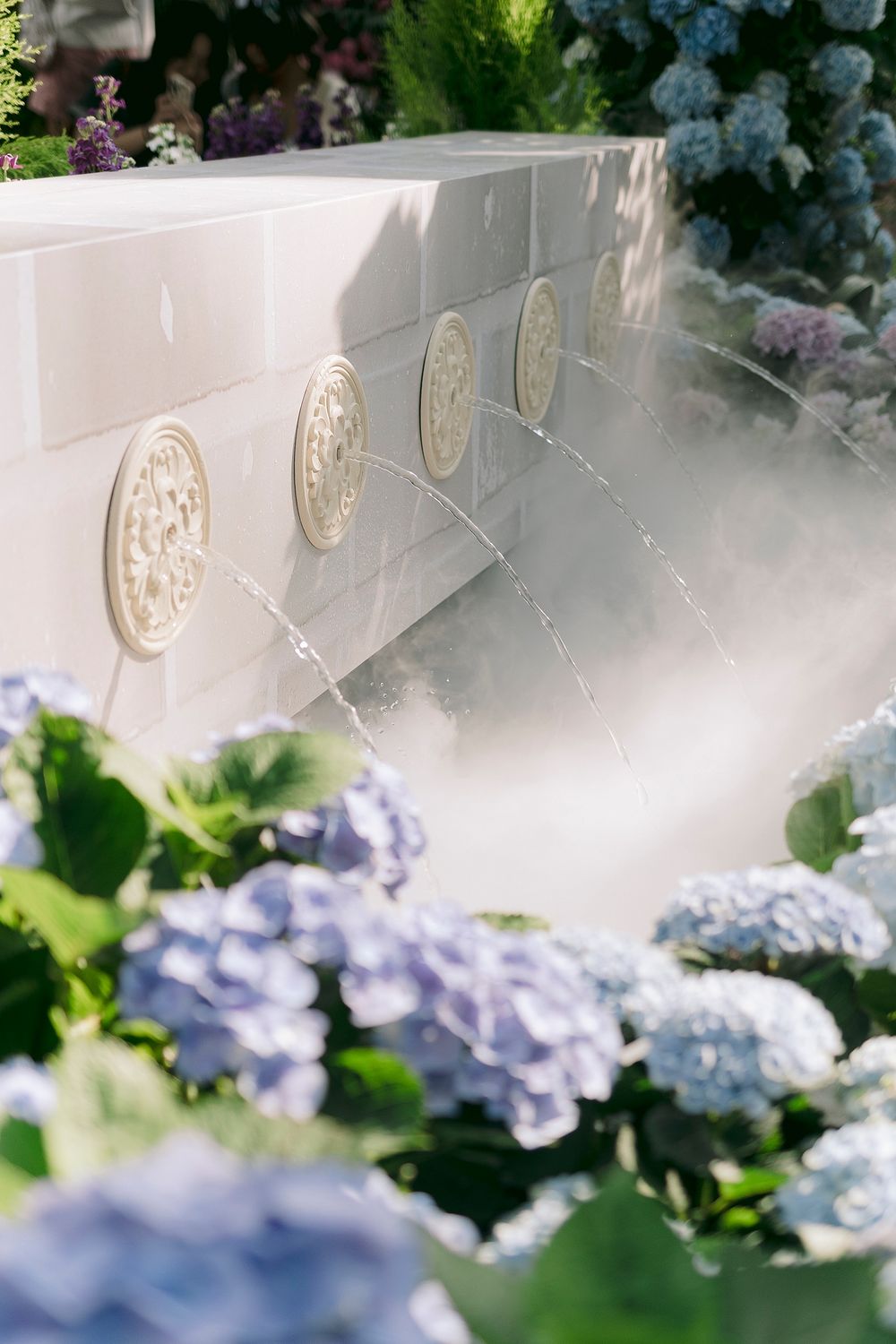 White stone fountain wall with ornate medallions surrounded by blooming blue hydrangeas in a sunlit garden.