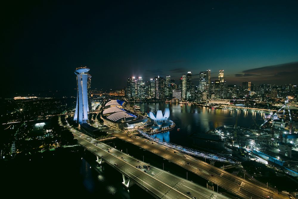 Aerial night view of Singapore skyline with Marina Bay Sands, glowing city lights, and illuminated highways reflecting on the water.