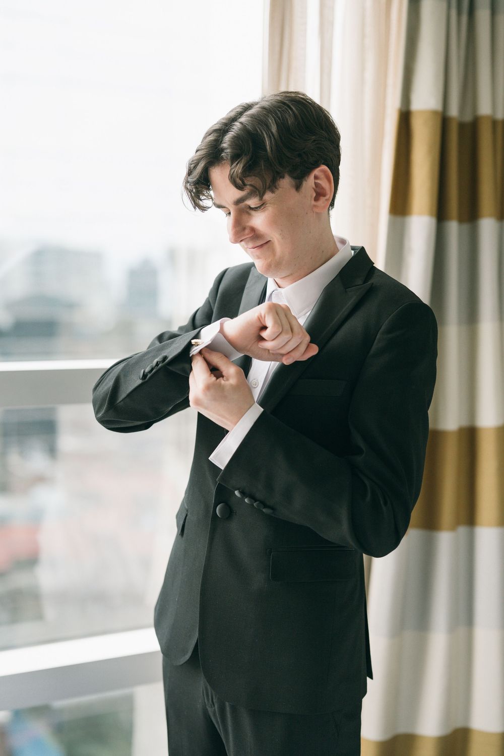Young man in black suit adjusting cufflinks by window, smiling, dressed for formal event.