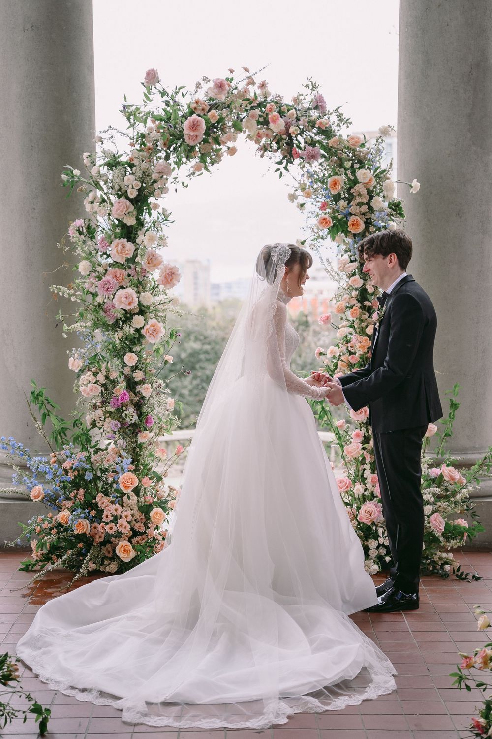 Bride and groom exchange vows under a lush floral arch adorned with pink and white roses at an elegant outdoor wedding.