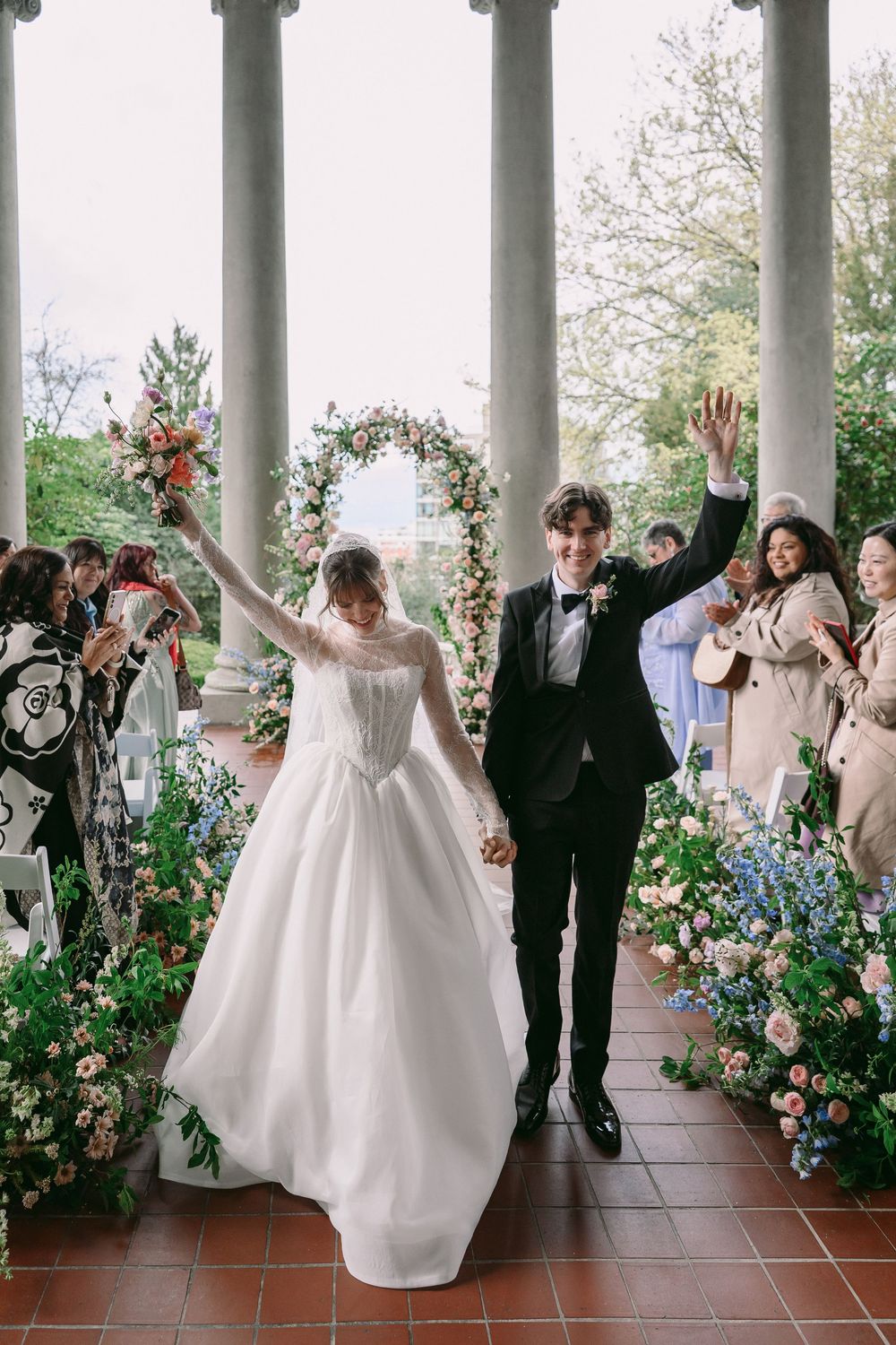Joyful newlyweds raise their hands walking back down the aisle past guests and floral arrangements after their ceremony.