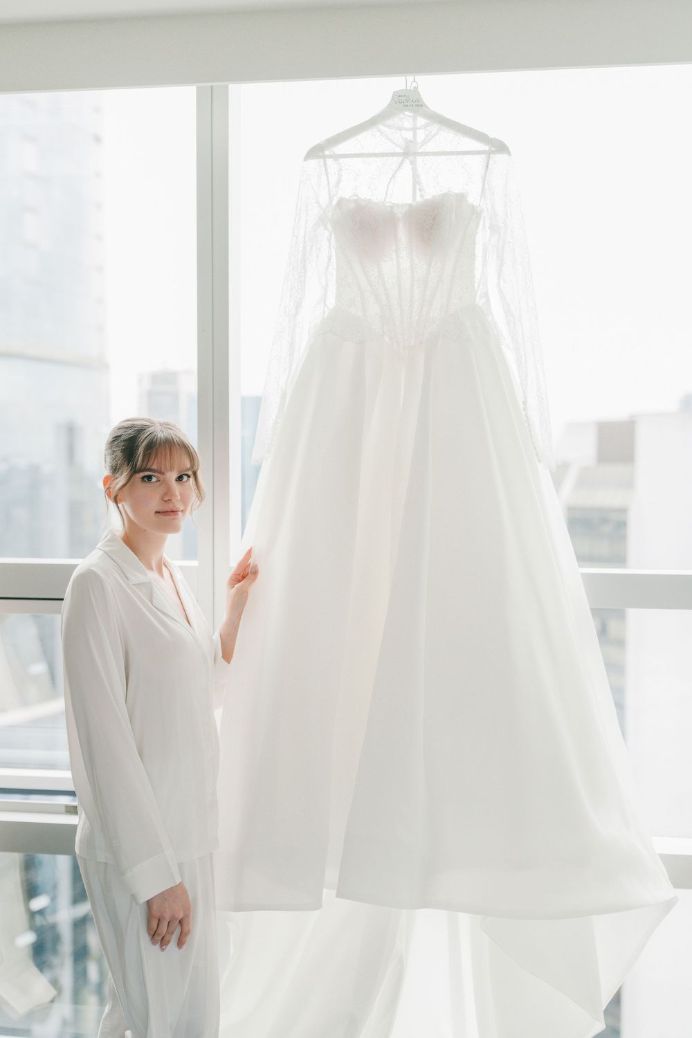 Bride in white robe admiring her elegant strapless ball gown wedding dress hanging by a bright window.