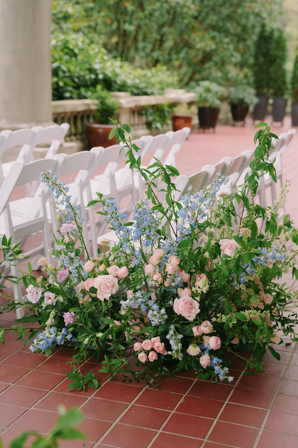 Lush garden wedding ceremony aisle with pink roses and blue delphinium floral arrangements on red brick terrace.