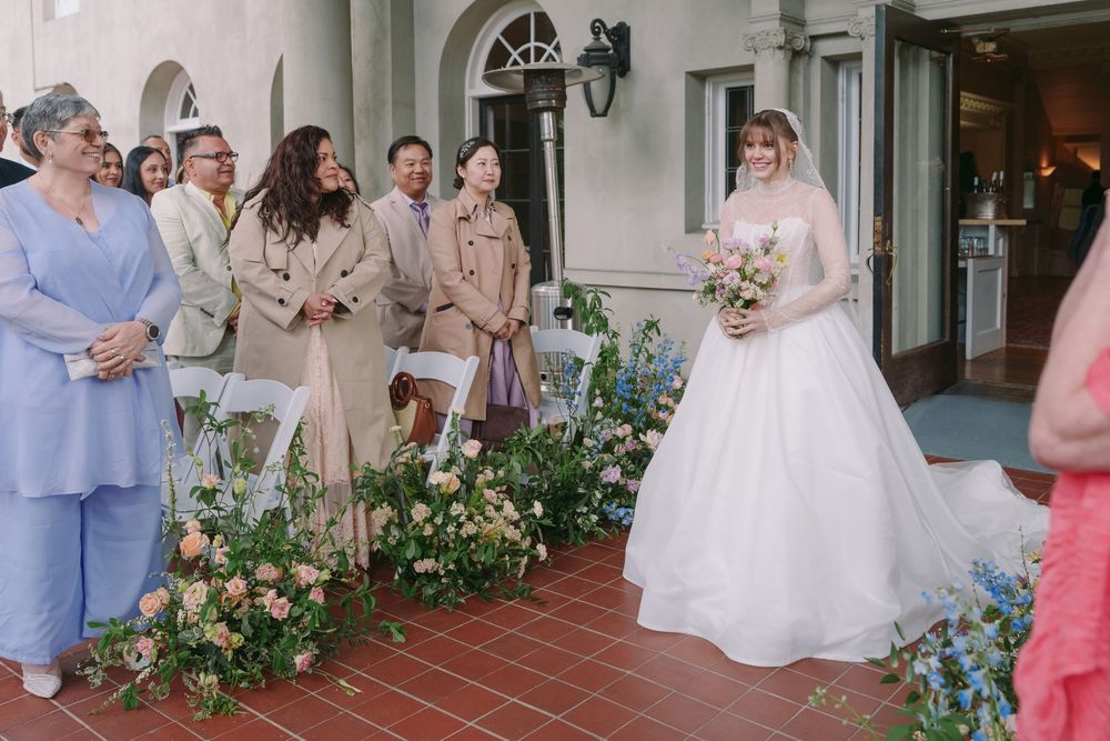 Bride in white ball gown holding bouquet walks down aisle past smiling guests at outdoor wedding ceremony.