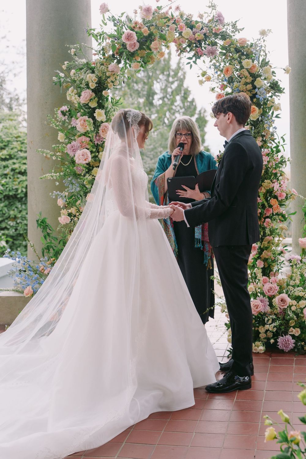 Bride and groom exchange vows beneath a lush floral arch with pink roses, officiated outdoors on a brick terrace.