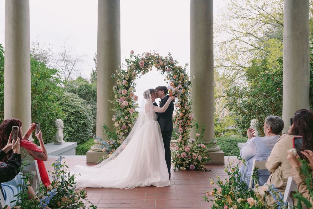 Bride and groom kissing under a floral arch at an outdoor wedding ceremony with elegant stone columns.