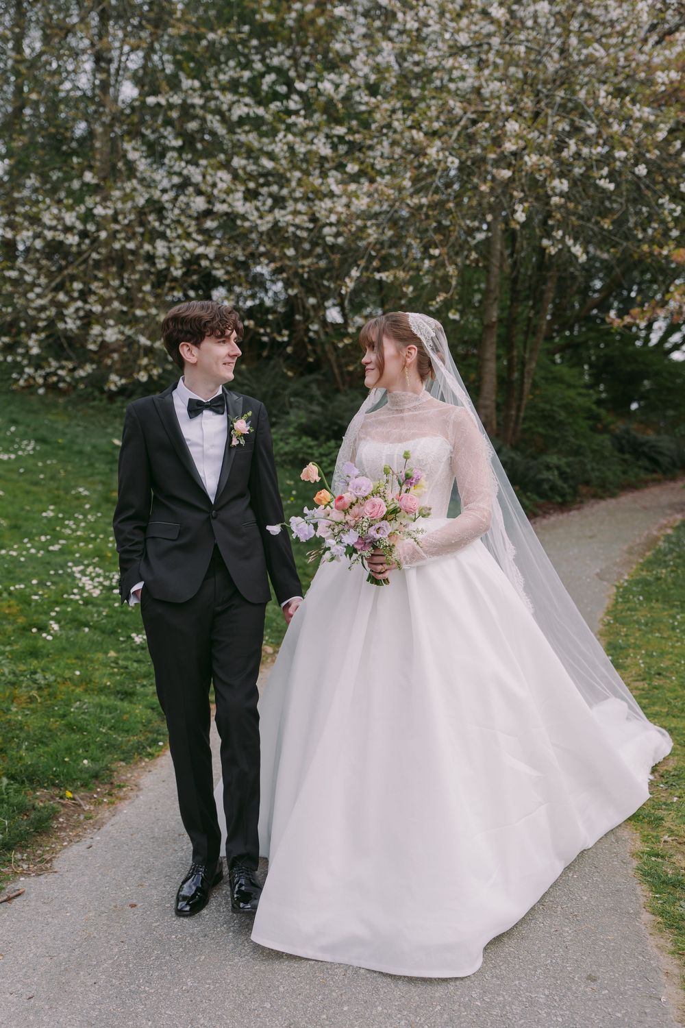 Bride and groom walking hand-in-hand on a garden path with blooming white trees in spring.
