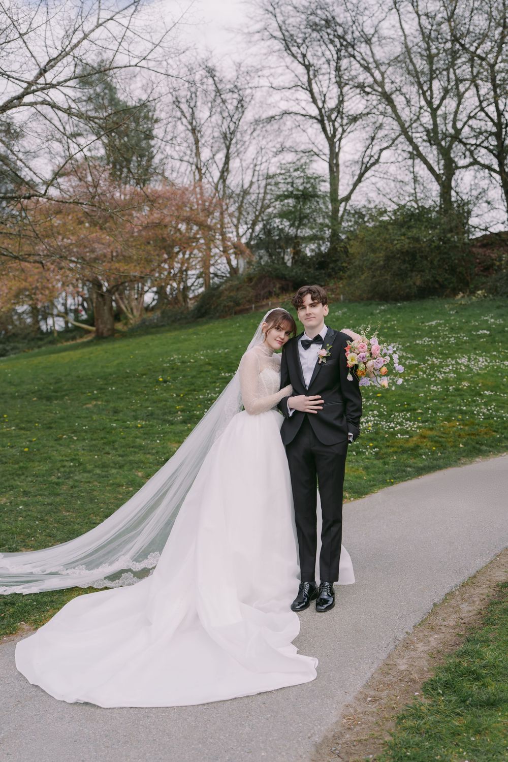 Bride and groom pose on garden path, she in white gown with long veil, he in black tuxedo with bow tie.