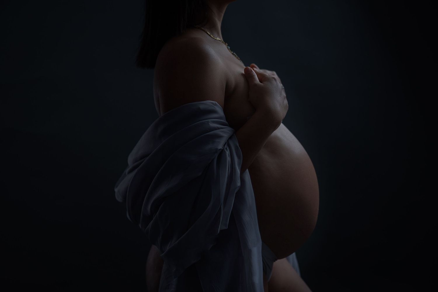 Artistic maternity photo of pregnant woman in dramatic low-key lighting, draped in blue fabric, cradling her bare belly.