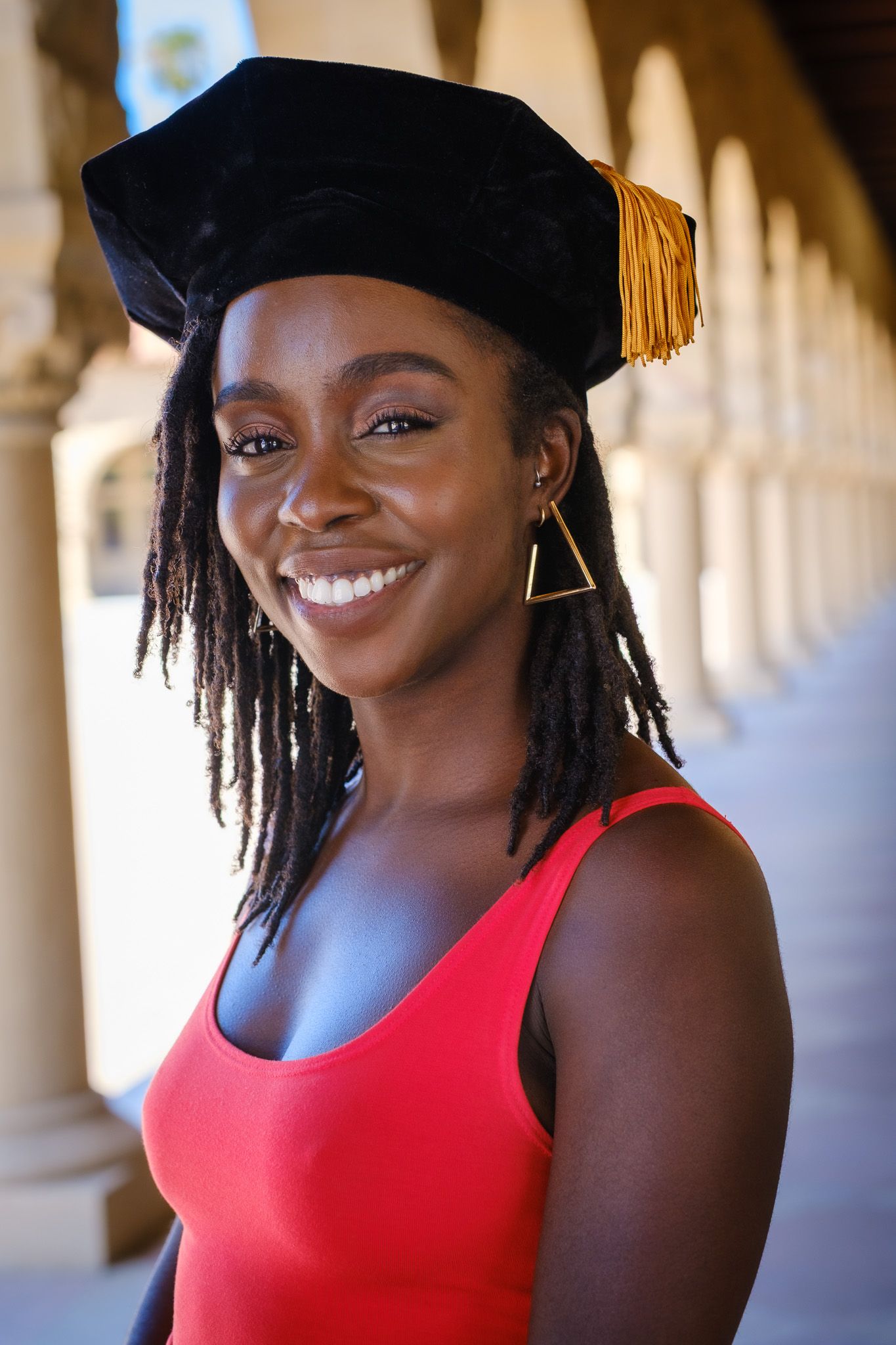 Smiling young Black woman wearing a doctoral graduation cap with gold tassel and red dress, standing in an archway.