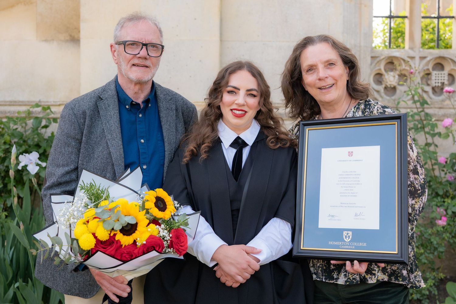 Three people pose outdoors at a graduation ceremony, holding flowers and a framed certificate.